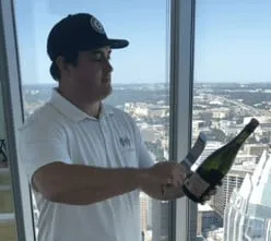 A man in a cap and polo shirt opening a bottle of champagne by a high-rise office window overlooking the city